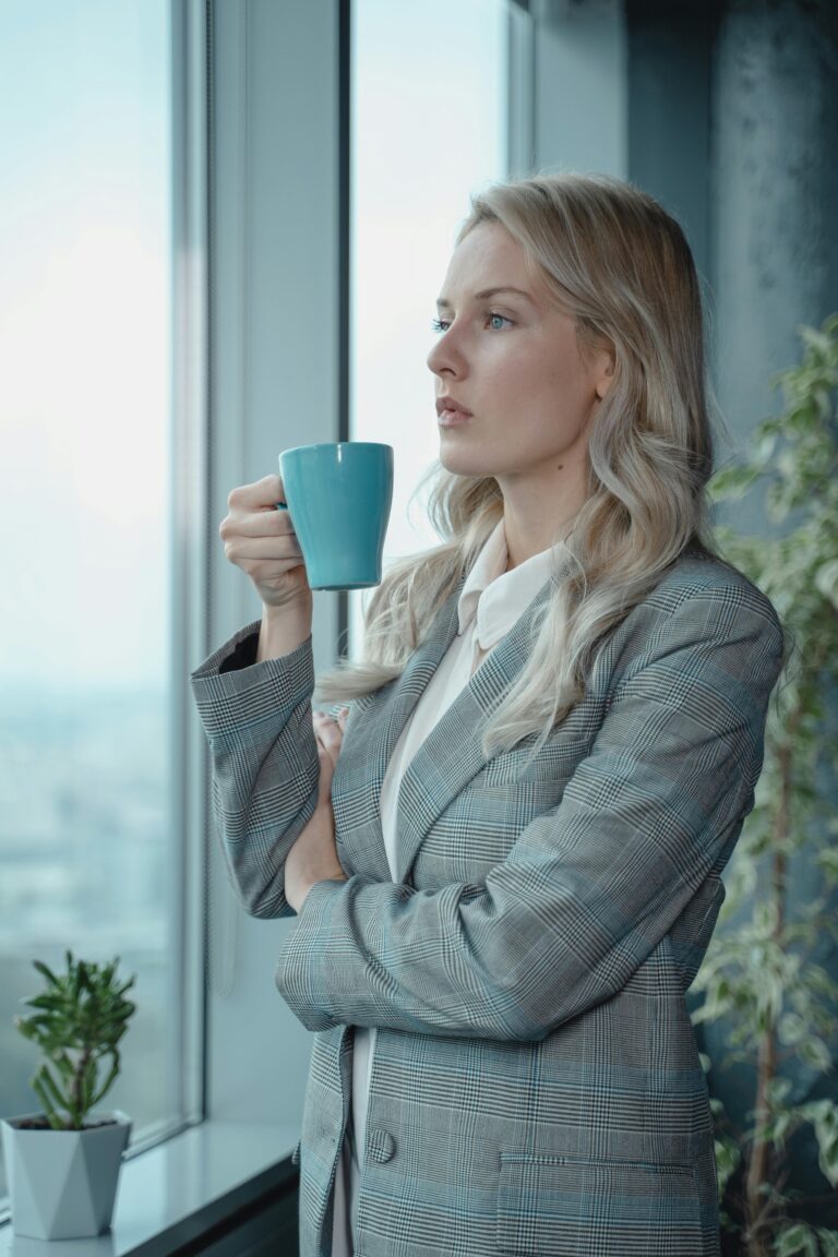 A focused businesswoman in a suit holding coffee, gazing outside the office window.